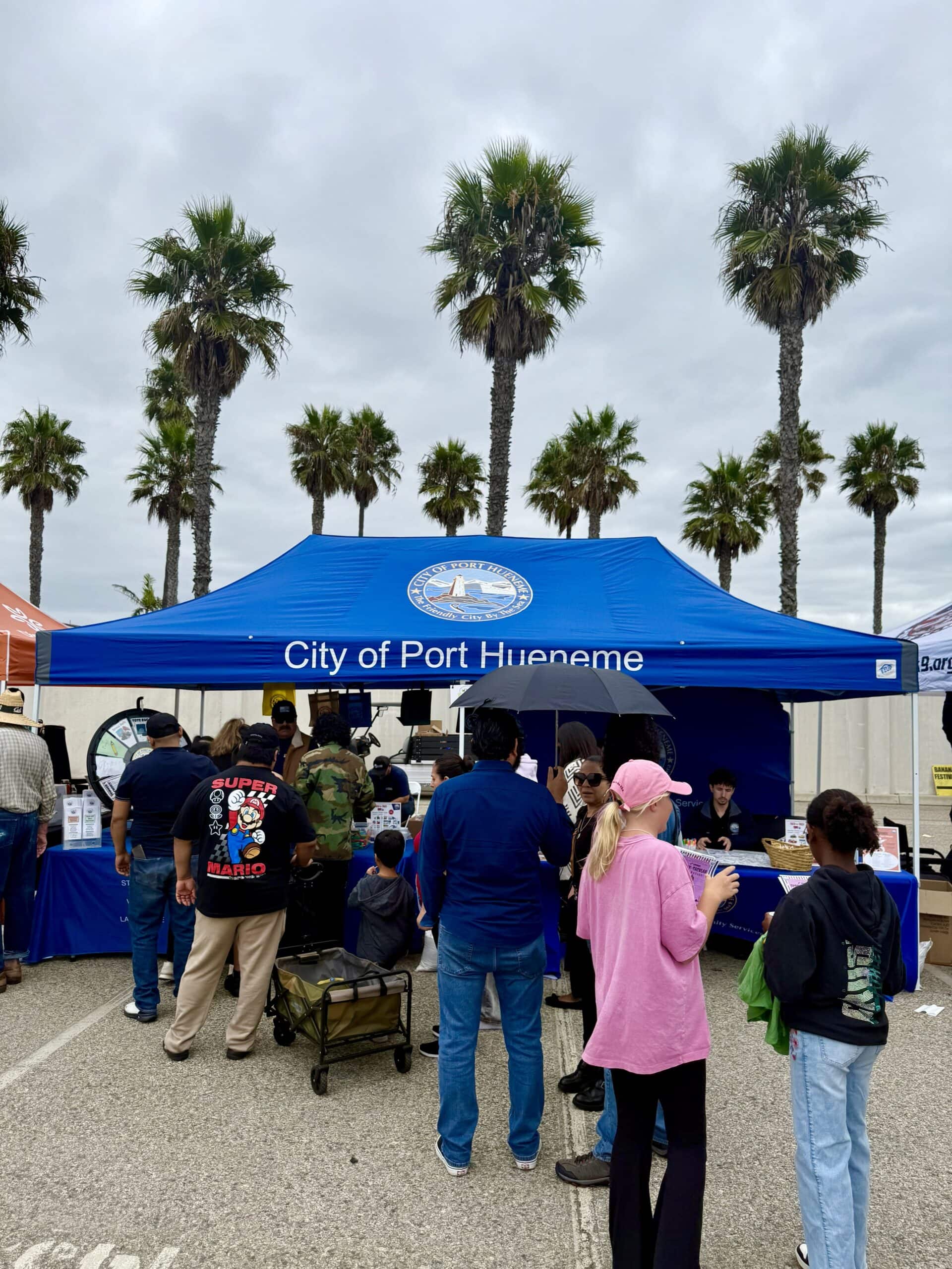 Banana Festival in Port Hueneme Crowd at Banana Festival under blue tent with palm trees in background.
